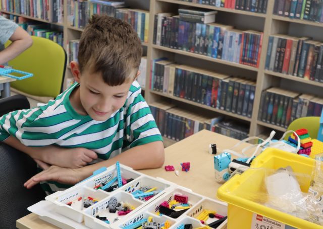 Na zdjęciu znajduje się chłopiec przy stoliku, na którym leżą klocki LEGO, z których układa samochód. W tle znajdują się półki z książkami.