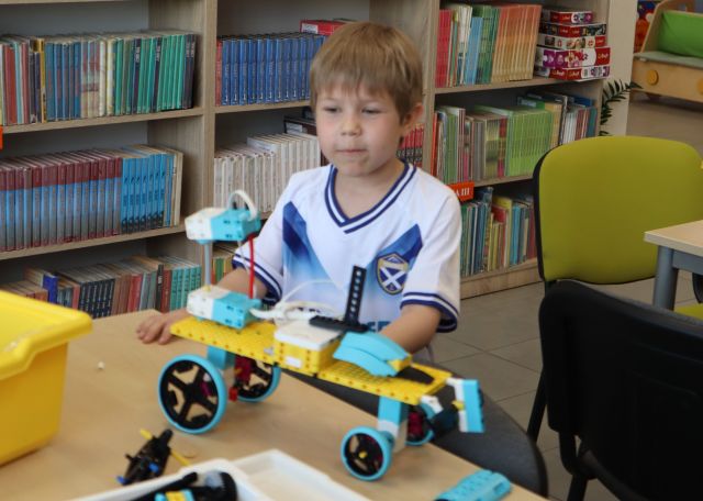 Na zdjęciu znajduje się chłopiec stojący przy stoliku, na którym leży zbudowany z klocków LEGO pojazd. W tle znajdują się półki z książkami.