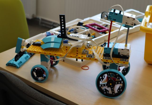 Na zdjęciu znajduje się pojazd zbudowany z klocków LEGO, który stoi na stole, a w tle znajdują się klocki LEGO w pudełku.