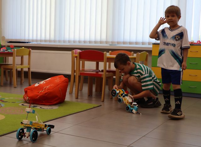 Na zdjęciu znajduje się dwóch chłopców, jeden kuca i poprawia swojego robota z klocków LEGO, a drugi stoi obok. W tle znajdują się małe stoliki z krzesełkami, po lewej stronie znajduje się dywan i czerwona pufa. A po prawej stronie przy dywanie stoi samochód zbudowany z klocków LEGO.