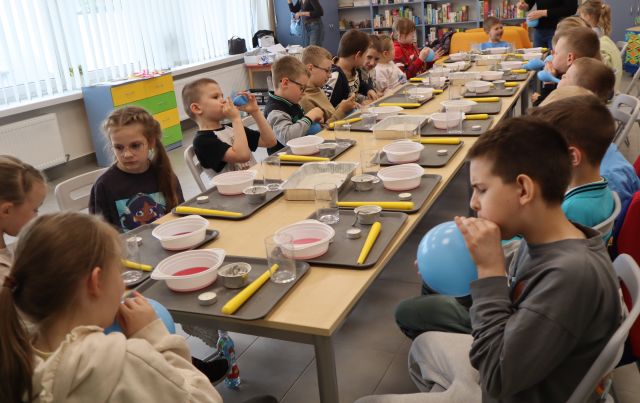 Na zdjęciu znajdują się siedzące przy złączonych stołach dzieci, które dmuchają balony. Na stolikach przed nimi leżą tacki ze świeczkami, z miską z wodą i szklanką. W tle znajdują się półki z grami oraz okna.