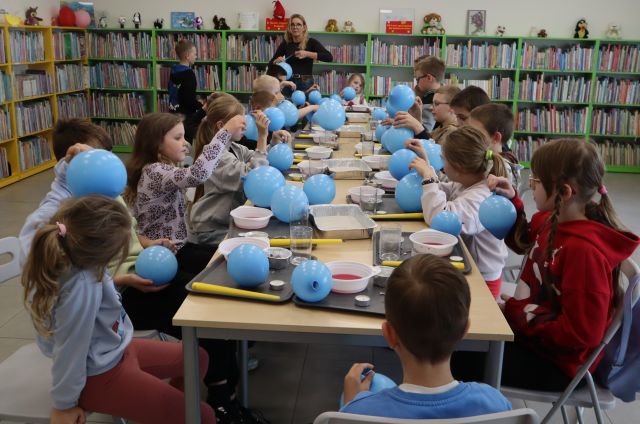 Na zdjęciu znajdują się dzieci siedzące przy stole, mają przed sobą balony. W tle znajduję się kobieta oraz półki z książkami.