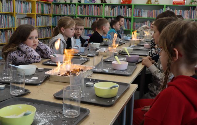 Na zdjęciu znajdują się dzieci przy złączonych stołach, przed sobą mają tacki z miskami i szklankami a na środku stoją aluminiowe tacki, w których wykonywane są węże faraona. W tle znajdują się półki z książkami.