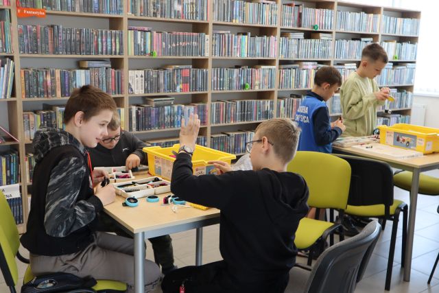 Na zdjęciu znajduje się piątka chłopców przy dwóch stolikach, przy których układają klocki LEGO. W tle znajdują się półki z książkami.