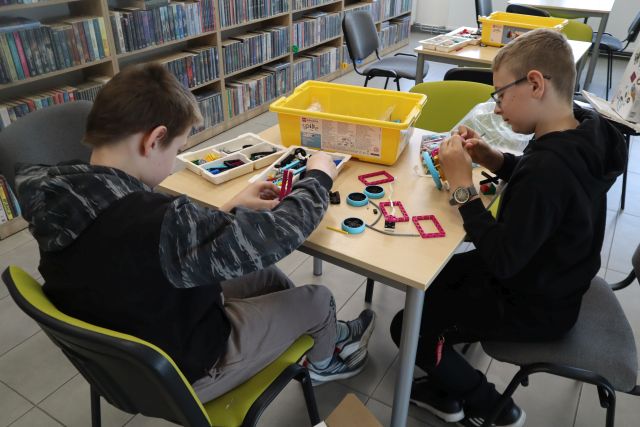 Na zdjęciu znajduje się dwóch chłopców, którzy składają klocki LEGO przy stoliku. W tle znajdują się półki z książkami oraz stoliki.