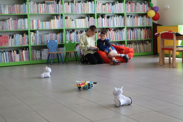 Na zdjęciu znajduje się dwóch chłopców siedzących na pufie i za pomocą tabletu sterują robotem LEGO, obok niego znajdują się jeszcze dwa roboty PHOTON. W tle znajdują się półki z książkami, dwa krzesłka.