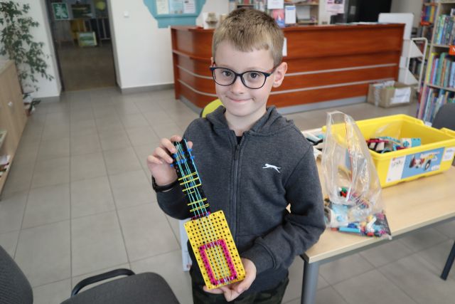 Na zdjęciu znajduje się chłopiec, który trzyma w ręce zbudowaną z klocków LEGO gitarę, za nim stoi stół z klockami oraz znajduje się biurko.