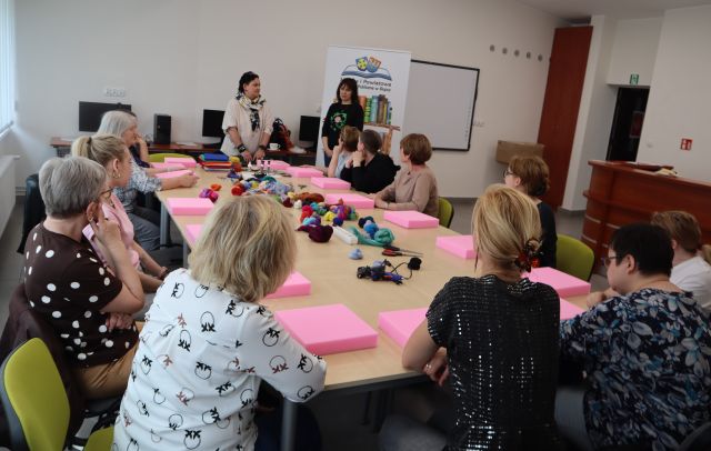 Na zdjęciu znajdują się kobiety siedzące przy złączonych stołach, przed sobą mają różowe kwadratowe gąbki, a na środku stołu leży wełna czesana. Przy stołach stoją dwie kobiety. Warsztaty odbywają się w sali komputerowej.
