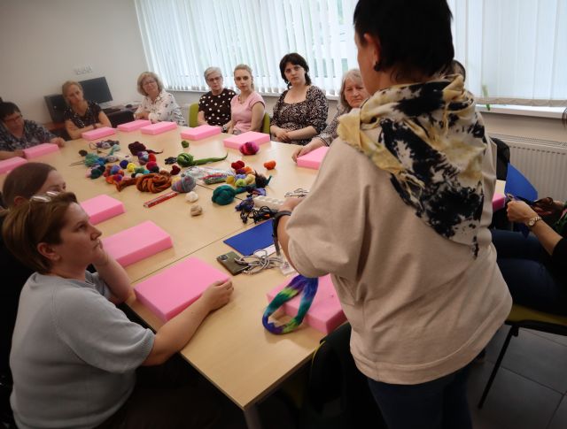 Na zdjęciu znajdują się kobiety przy złączonych stołach, przed sobą mają różowe gąbki oraz wełne. Warsztaty odbywają się w sali komputerowej.