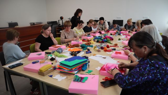 Na zdjęciu znajdują się kobiety przy złączonych stołach, które filcują wycięte przez siebie wzory na różowych gąbkach. Na środku stołu leży wełna czesana, filc. Warsztaty odbywają się w sali komputerowej.