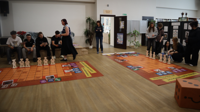 Na zdjęciu znajduje się młodzież przy dwóch planszach do gry wielkoformatowej, po prawej stronie znajduje się duża kostka do gry. Gra odbywa się na holu biblioteki.