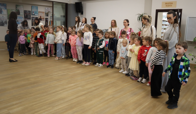 Na zdjęciu znajduje się około pięćdziesięciu przedszkolaków oraz oraz dziesięć kobiet, grupa stoi na holu biblioteki.
