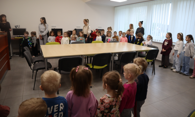 Na zdjęciu znajdują się dzieci wraz z pięcioma kobietami w sali komputerowej, na środku sali stoją złączone stoliki z krzesełkami.