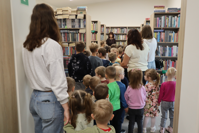 Na zdjęciu znajdują sie przedszkolaki wraz z czterema kobietami i idą między regałami z książkami w bibliotece. Zdjęcie zostało wykonane od tyłu.