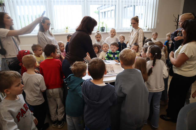 Na zdjęciu znajdują się przedszkolaki ustawione dookoła stolika przy którym kobieta pokazuje bardzo dużą książkę. Wraz z dziećmi stoją cztery kobiety. W tle znajdują się okna.