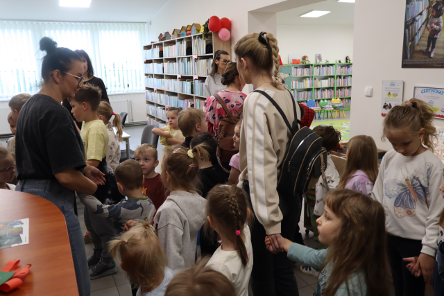 Na zdjęciu znajdują się dzieci wchodzące do sali bibliotecznej wraz z przedszkolankami. W tle znajdują się półki z książkami, a po lewej stronie zdjęcia znajduje się kawałek biurka.