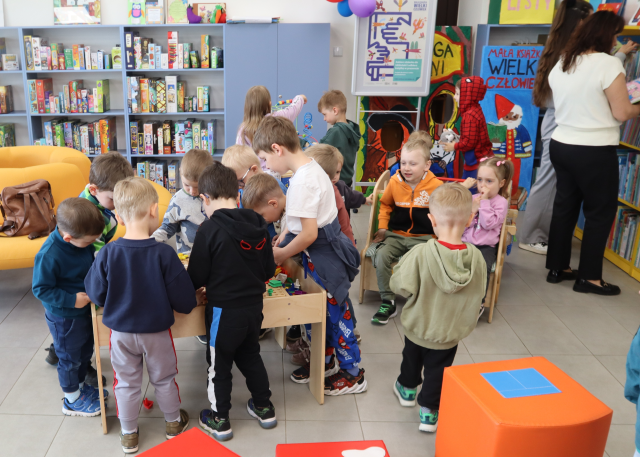Na zdjęciu znajdują się przedszkolaki bawiące się stolikiem do majsterkowania, samochodzikiem. W tle znajdują się półki z grami. Po prawej stronie stoi kobieta i przegląda znajdujące się na półce książki.