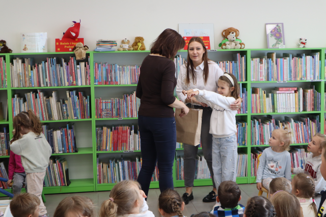 Na zdjęciu znajdują się dwie kobiety oraz dziewczynka, które daje torbę jednej z nich. W tle znajdują się półki z książkami, a przed kobietami siedzą dzieci.