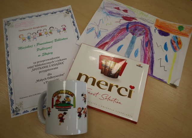 Na zdjęciu znajduje się podziękowanie za zajęcia, kolorowy rysunek, merci oraz kubek z logiem przedszkola. Upominki leżą na stoliku.