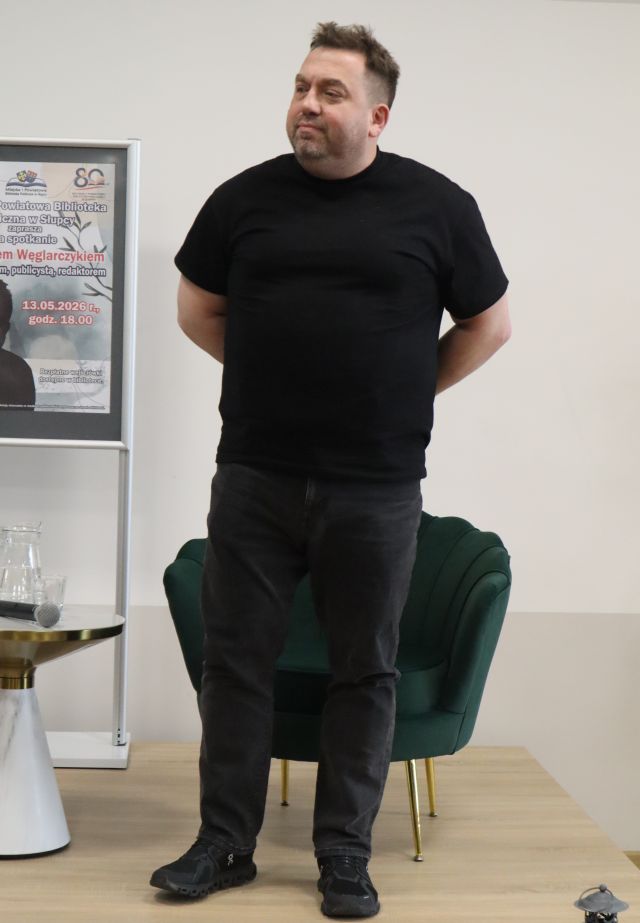 Na zdjęciu znajduje się stojący na scenie Bartosz Węglarczyk, ręce ma założone za plecami. Za mężczyzną stoi zielony fotel, a po lewej stronie stoi stolik, na którym znajduje się mikrofon oraz woda, za stolikiem stoi plakat promujący spotkanie.