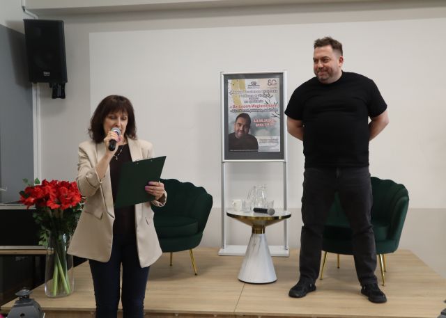 Na zdjęciu znajduje się stojący na scenie mężczyzna ze złożonymi za plecami rękoma, za mężczyzną znajduje się fotel, a po lewej stronie stolik z wodą i mikrofonem, plakat promujący spotkanie oraz jeszcze jeden fotel. Przed sceną stoi kobieta z mikrofonem oraz podkładką na kartkę w ręce, z której czyta, po jej lewej stronie stoją czerwone kwiaty w wazonie.