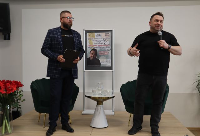 Na zdjęciu znajduje się dwóch mężczyzn stojących na scenie, mężczyzna stojący po prawej stronie mówi przez mikrofon. Za mężczyznami znajdują się zielone fotele, a pomiędzy nimi stolik z wodą oraz plakat promujący spotkanie. Po lewej stronie na rogu sceny stoją czerwone kwiaty.