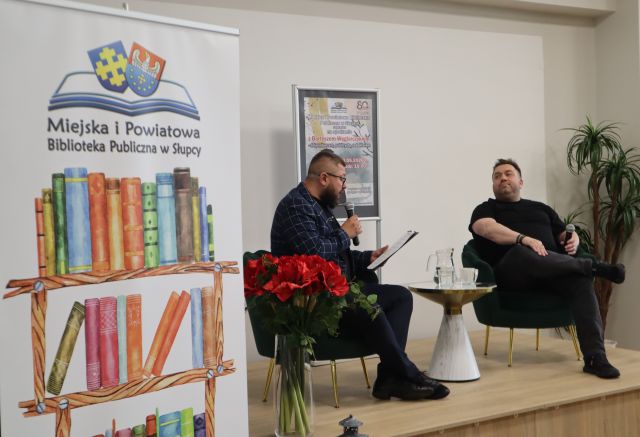 Na zdjęciu znajduje się dwóch mężczyzn siedzących na scenie, pomiędzy nimi znajduje się stolik z wodą oraz plakat promujący spotkanie. W centrum po lewej stronie zdjęcia znajduje się baner z logiem biblioteki oraz książkami.