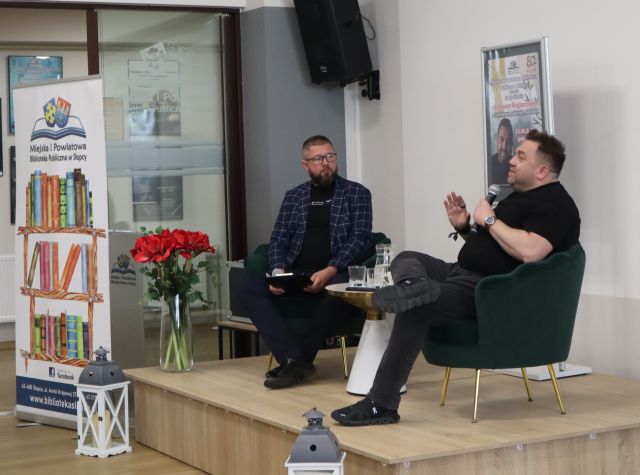 Na zdjęciu zrobionym od boku znajduje się dwóch mężczyzn na scenie, którzy siedzą na fotelach i jeden mówi przez mikrofon. Pomiędzy nimi znajduje się stolik z wodą oraz plakat promujący spotkanie. Na scenie po lewej stronie na rogu stoją czerwone kwiaty, pod sceną znajdują się dwa lampiony oraz baner z logiem biblioteki i książkami po lewej stronie. Na ścianach w tle wiszą plakaty.