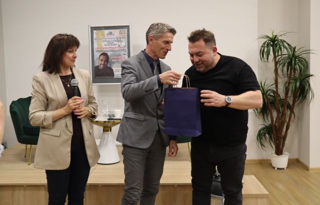 Na zdjęciu znajduje się dwóch mężczyzn, jeden podaje drugiemu granatową torbę z upominkiem, po lewej stronie stoi kobieta z mikrofonem. W tle znajduje się scena z fotelami, stolikiem i plakatem promującym spotkanie. Przy scenie po prawej stronie stoi roślina.