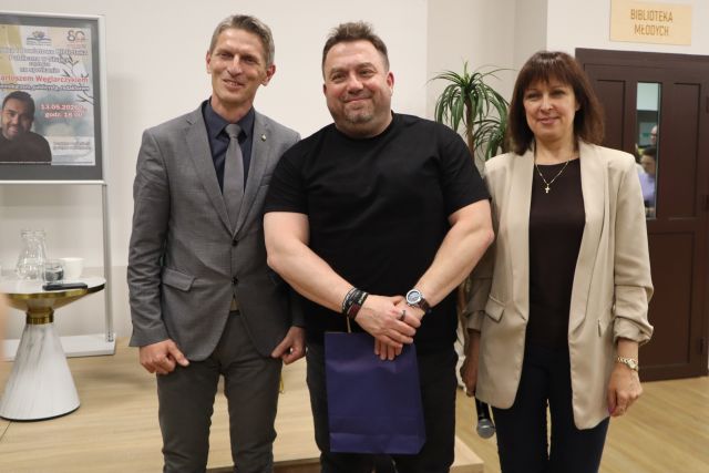 Na zdjęciu znajduje się trójka osób, dwóch mężczyzn oraz kobieta po prawej, mężczyzna stojący na środku trzyma w ręce upominek w granatowej torbie. W tle znajdują się drzwi do sali bibliotecznej, a po lewej stronie scena ze stolikiem i plakatem promującym spotkanie.