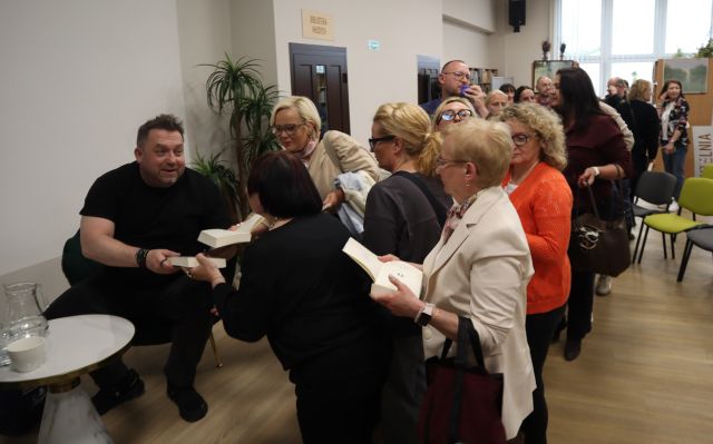 Na zdjęciu znajduje się tłum ludzi, w kolejce po autograf od Bartosza Węglarczyka, który siedzi na scenie, przy nim stoi stolik.