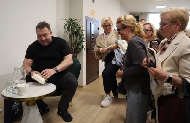 Na zdjęciu znajdują się kobiety stojące przed sceną, na której siedzi mężczyzna i podpisuje książkę na stoliku z wodą. W tle znajduję się roślina, drzwi do sali bibliotecznej.