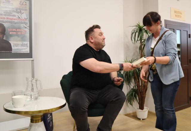 Na zdjęciu znajduje się mężczyzna siedzący na scenie, który podaje książkę kobiecie stojącej po prawej stronie. Za kobietą w tle znajdują się drzwi oraz kwiat, a po lewej stronie zdjęcia znajduje się stolik z wodą oraz plakat promujący spotkanie.