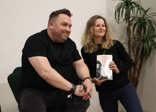 Na zdjęciu znajduje się siedzący na fotelu, na scenie mężczyzna, po lewej stronie stoi kobieta z jego książką do zdjęcia. Za kobietą znajduje się wysoka rośłina.