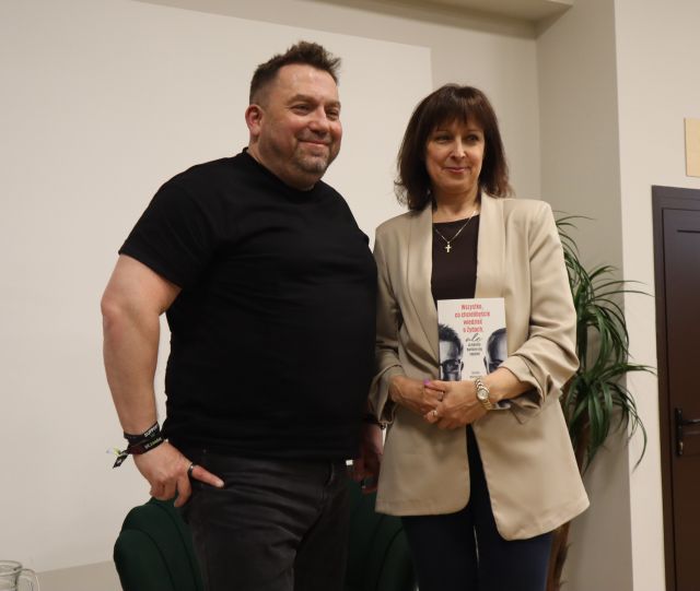 Na zdjęciu znajduje się kobieta, która trzyma książkę i stoi do zdjęcia z Bartoszem Węglarczykiem, za nimi znajduje się kwiat oraz fotel.