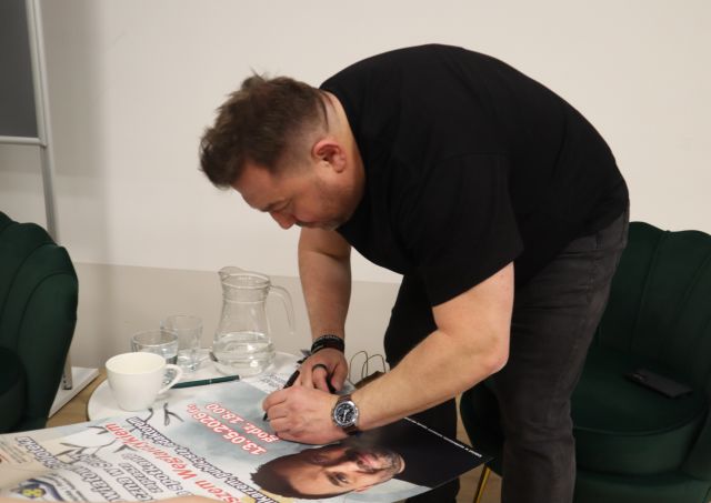 Na zdjęciu znajduje się mężczyzna nachylający się nad leżącym na stoliku plakatem i go podpisuje, na stoliku leżą również szklanki i dzbanek.