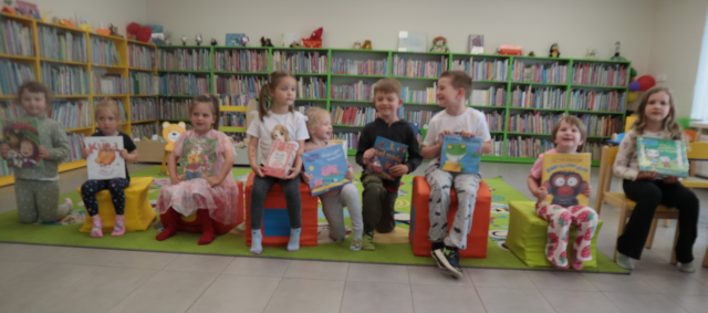 Na zdjęciu znajduje się dziewięcioro dzieci, którzy siedzą na pociągu z puf. W tle znajduje się dywan i półki z książkami. 