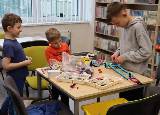 Na zdjęciu znajduje się trójka chłopców, którzy układają klocki LEGO. W tle znajduje się jeszcze jeden stół, półki z książkami oraz okna.