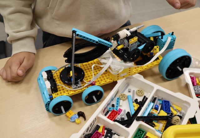 Na zdjęciu znajduje się wykonany z klocków LEGO samochód leżący na stole koło klocków, przy stoliku stoi chłopiec, którego ręce są widoczne na zdjęciu.