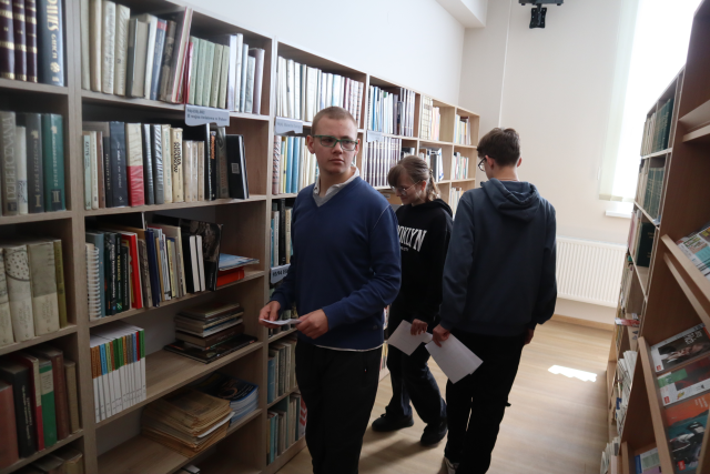 Na zdjęciu znajdują się trzy osoby między półkami w bibliotece, w ręce trzymają kartkę.