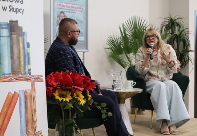 Na zdjęciu znajduje się kobieta wraz z mężczyzną na scenie, kobieta ma mikrofon przy buzi, za nią znajdują się zielone rośliny. Po prawej stronie w centrum znajduje się fragment banera z książkami oraz czerwone i żółte kwiaty. Zdjęcie zostało zrobione od boku.