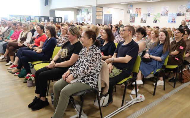 Na zdjęciu znajduje się widownia na holu biblioteki, ponad dwieście osób. W tle znajdują się okna, plakaty powieszone na ścianach.