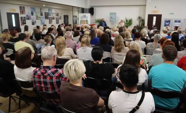 Na zdjęciu znajduje się widownia na holu biblioteki, przed nimi na scenie siedzi kobieta z mężczyzną za nimi stoi plakat promujący spotkanie, po lewej stronie przy scenie stoi baner z logiem biblioteki i książkami. W tle na ścianach znajdują się półki z książkami.
