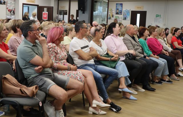 Na zdjęciu znajduje się widownia na holu biblioteki, w tle znajduje się przeszkolona ścianka i plakaty powieszone na ścianach.