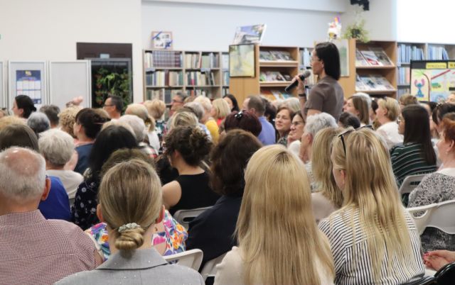 Na zdjęciu znajduje się fragment widowni na holu biblioteki, jedna kobieta stoi i mówi przez mikrofon. W tle znajdują się półki z książkami.