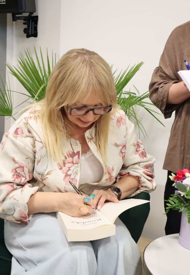 Na zdjęciu znajduje się kobieta, które podpisuje książkę, obok niej na stoliku stoją kwiaty oraz stoi kobieta, która nie została uchwycona cała na zdjęciu.