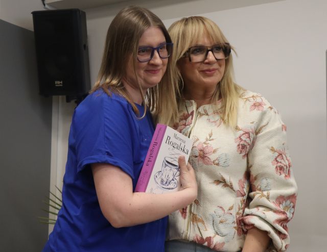 Na zdjęciu znajdują się dwie kobiety stojące do wspólnego zdjęcia, jedna z nich trzyma książkę. W tle znajduje się głośnik.