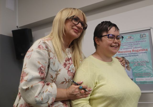 Na zdjęciu znajdują się dwie kobiety stojące do wspólnego zdjęcia. W tle znajduje się głośnik wiszący na ścianie oraz plakat po prawej stronie.