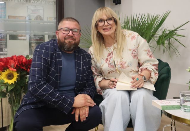 Na zdjęciu znajduje się siedząca kobieta i podpisująca książkę, obok niej kuca mężczyzna. W tle znajdują się kwiaty.
