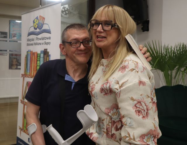 Na zdjęciu znajduje się kobieta i mężczyzna stoją do wspólnego zdjęcia. W tle znajduje się baner z książkami i logiem biblioteki oraz zielone drzewko.